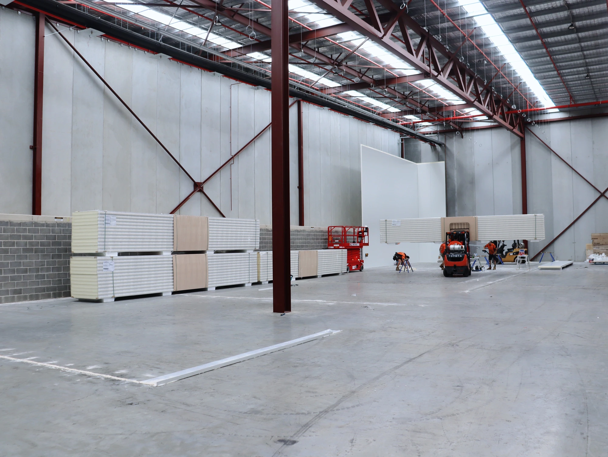The Chullora warehouse bay before the cool rooms went up, with insulated panel stacks landed and the first wall standing at the far end