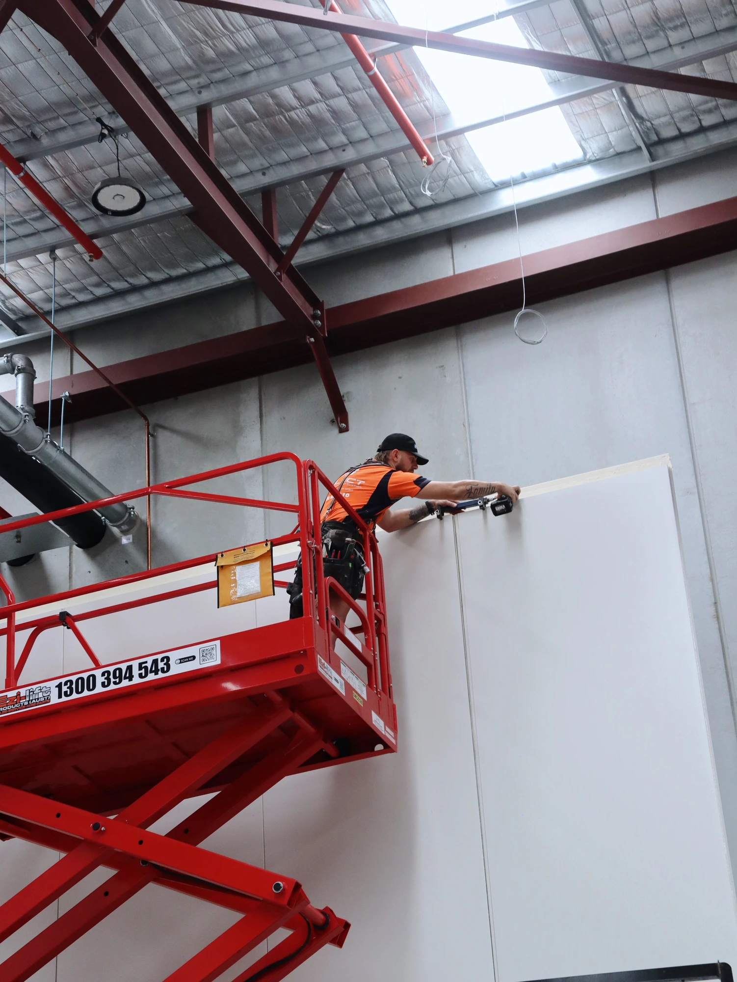 Installer fixing off the top of a full height wall panel from a scissor lift at Chullora, up in the warehouse roof structure